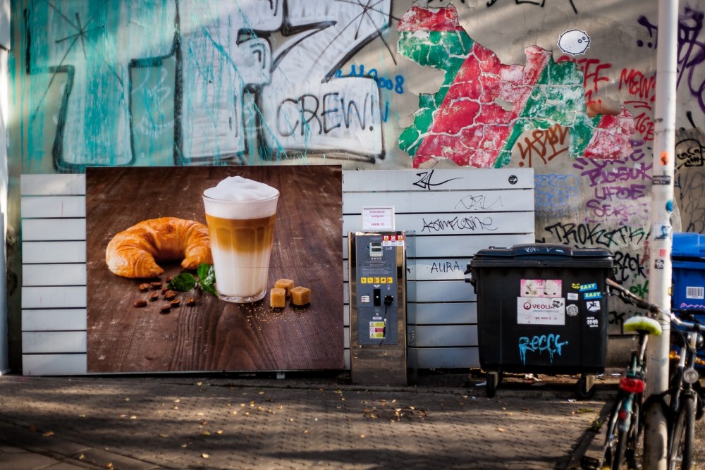 Ein Plakat mit einem Kaffee und Croissant auf einer Wand mit Graffitis und Mülleimern.