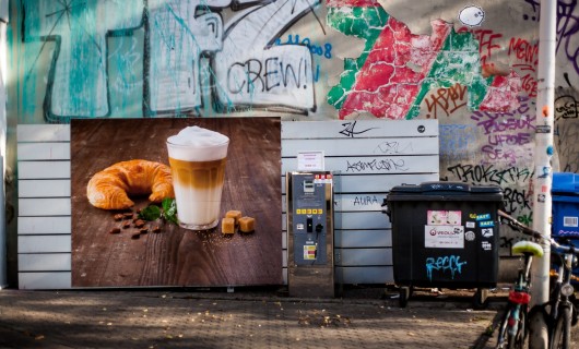 Ein Plakat mit einem Kaffee und Croissant auf einer Wand mit Graffitis und Mülleimern.