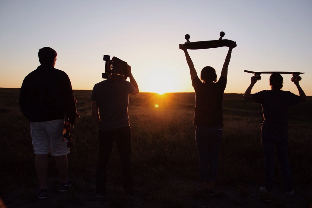 Vier Jugendliche sind von hinten zu sehen, wie sie in den Sonnenuntergang gehen und ihre Skateboards in der Hand halten.