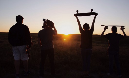Vier Jugendliche sind von hinten zu sehen, wie sie in den Sonnenuntergang gehen und ihre Skateboards in der Hand halten.