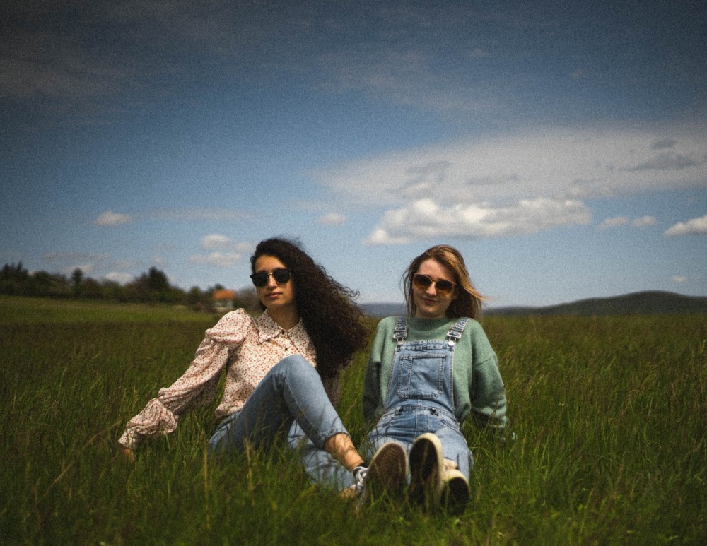 Zwei Frauen sitzen auf einer Wiese und schauen in die Kamera.