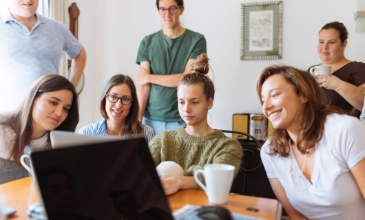 Sieben junge Menschen sitzen und stehen vor einem Laptop, auf den sie gemeinsam schauen. 