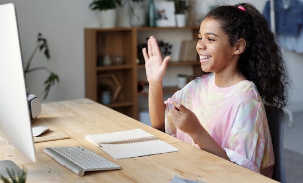 Mädchen sitzt lachend vor dem Computer und hebt die rechte Hand