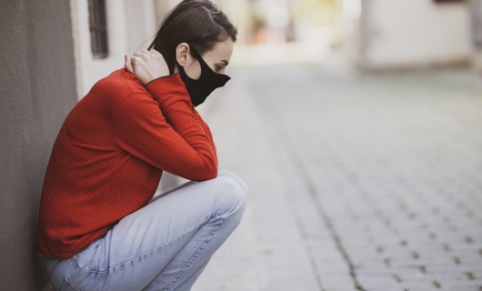 Eine Frau sitzt in der Hocke, die Hände im Nacken verschränkt mit einer Mund-Nasen-Maske.